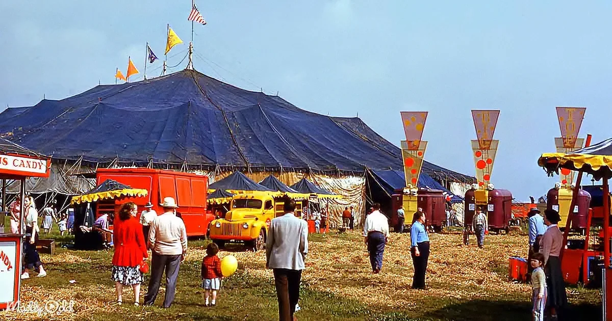 A large blue circus tent with people and vintage vehicles around it on a sunny day.