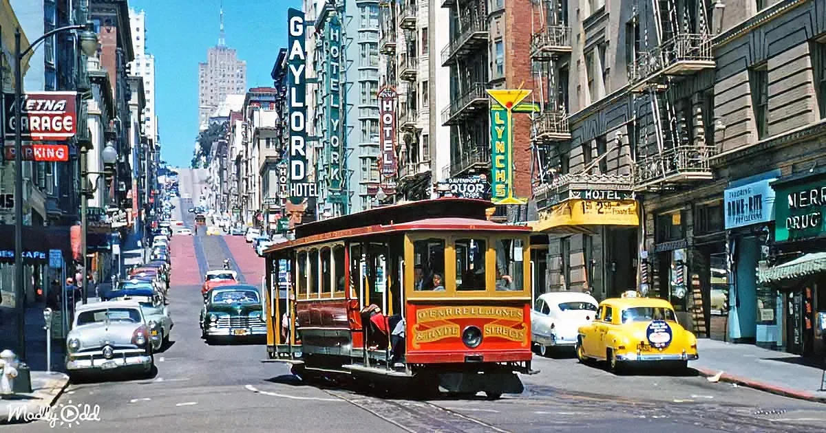 A vintage cable car climbs steep San Francisco street with classic cars and colorful signs.