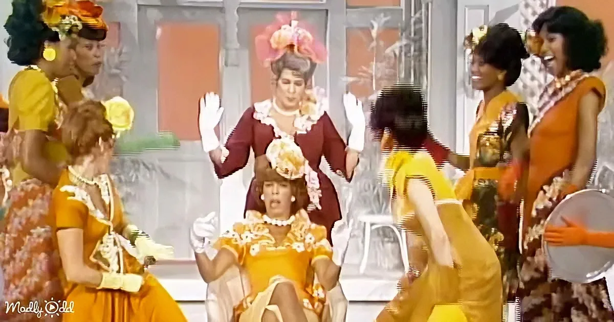 A woman in a red dress with white gloves stands center, surrounded by dancers in yellow and orange outfits.