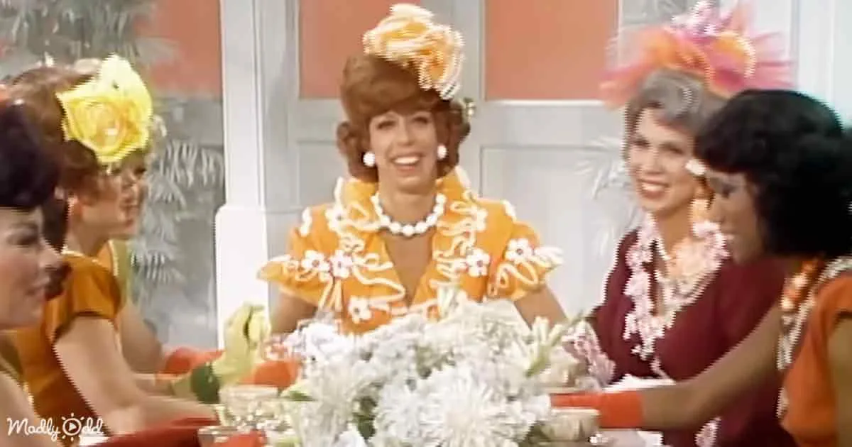A woman in a vibrant orange dress with floral details and a matching hat is seated at a table with others, smiling.