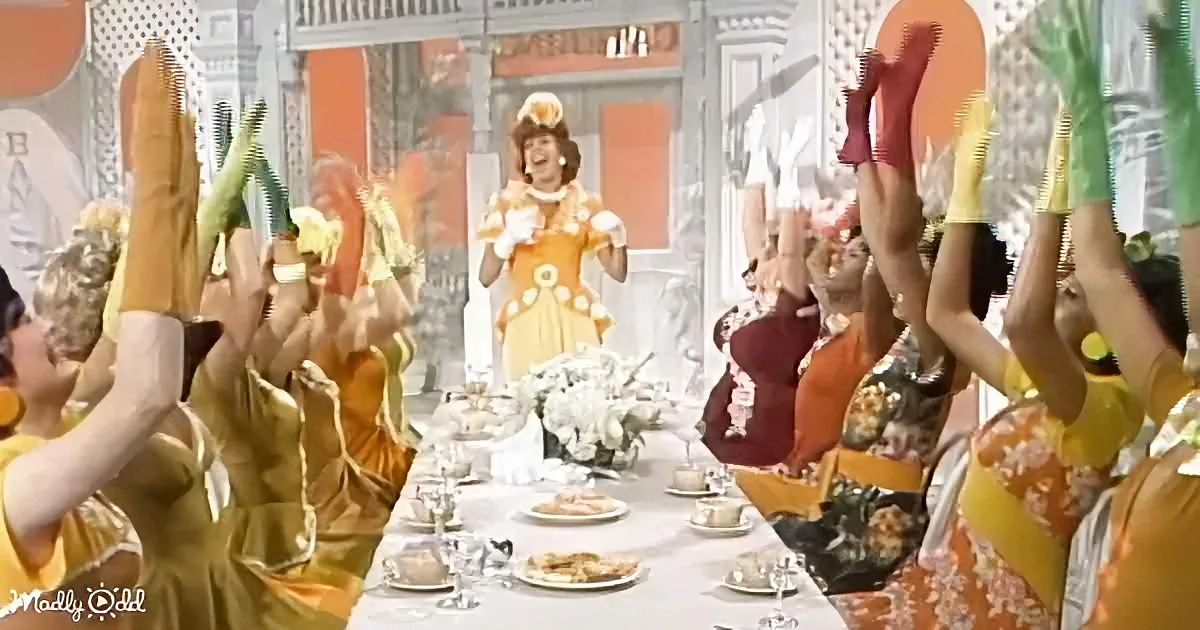 A woman in a yellow dress stands at a long table with people in yellow and floral dresses raising their hands.
