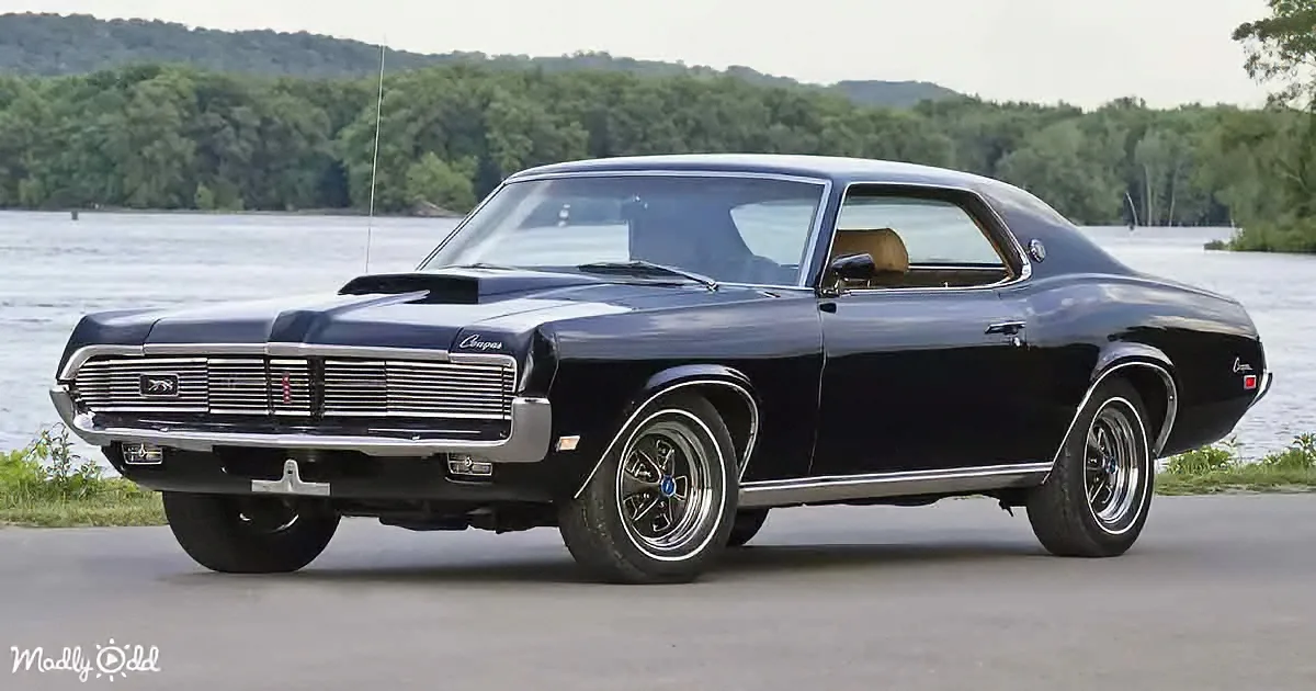 A classic black Mercury Cougar muscle car with chrome details and a sleek design is parked near a scenic lakeside.