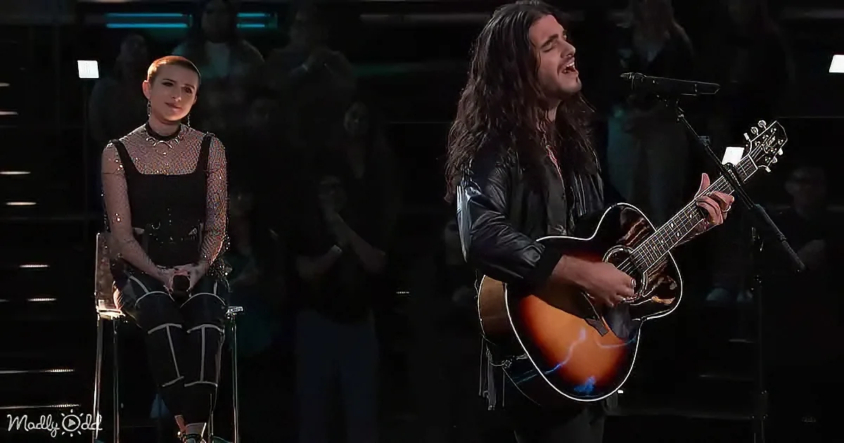 A man plays guitar and sings passionately while a woman in black attire sits calmly in the background.