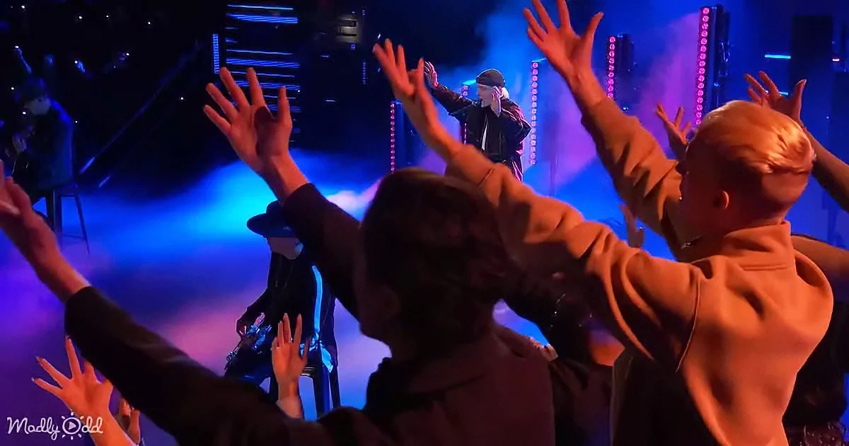 A performer on stage with raised arms, surrounded by an audience with hands up, under blue and purple stage lighting.