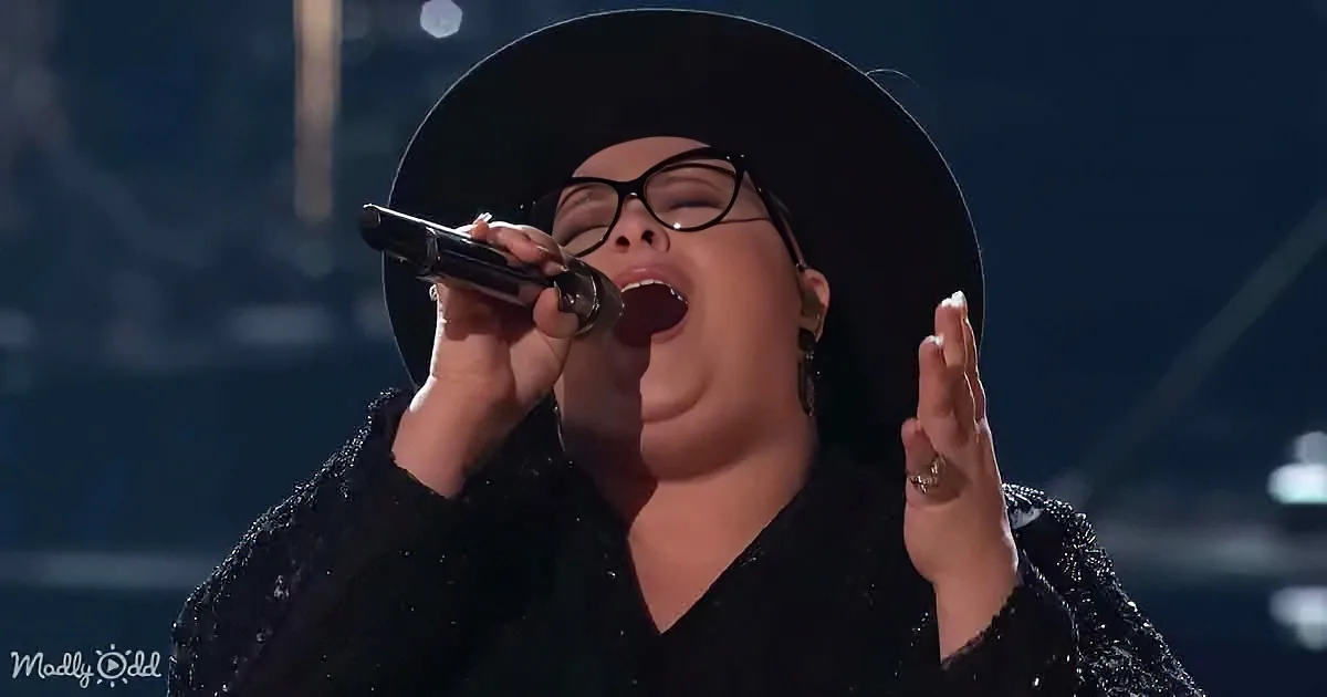 A person wearing a black hat and glasses sings passionately into a microphone, with one hand raised.