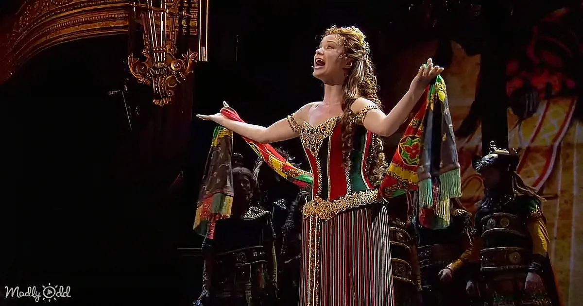 A performer in a vibrant, ornate costume holds colorful scarves, singing passionately on stage.