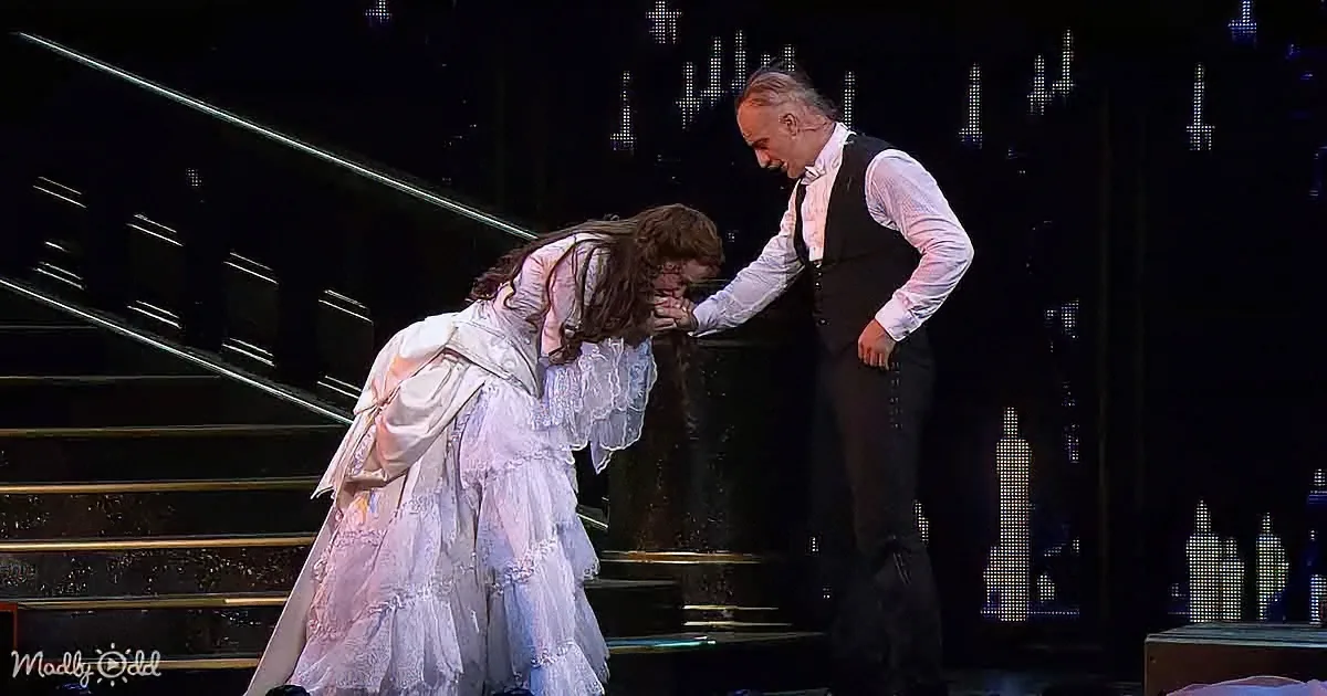 A woman in a white Victorian dress leans forward, hands clasped, while a man in a black vest and white shirt stands...