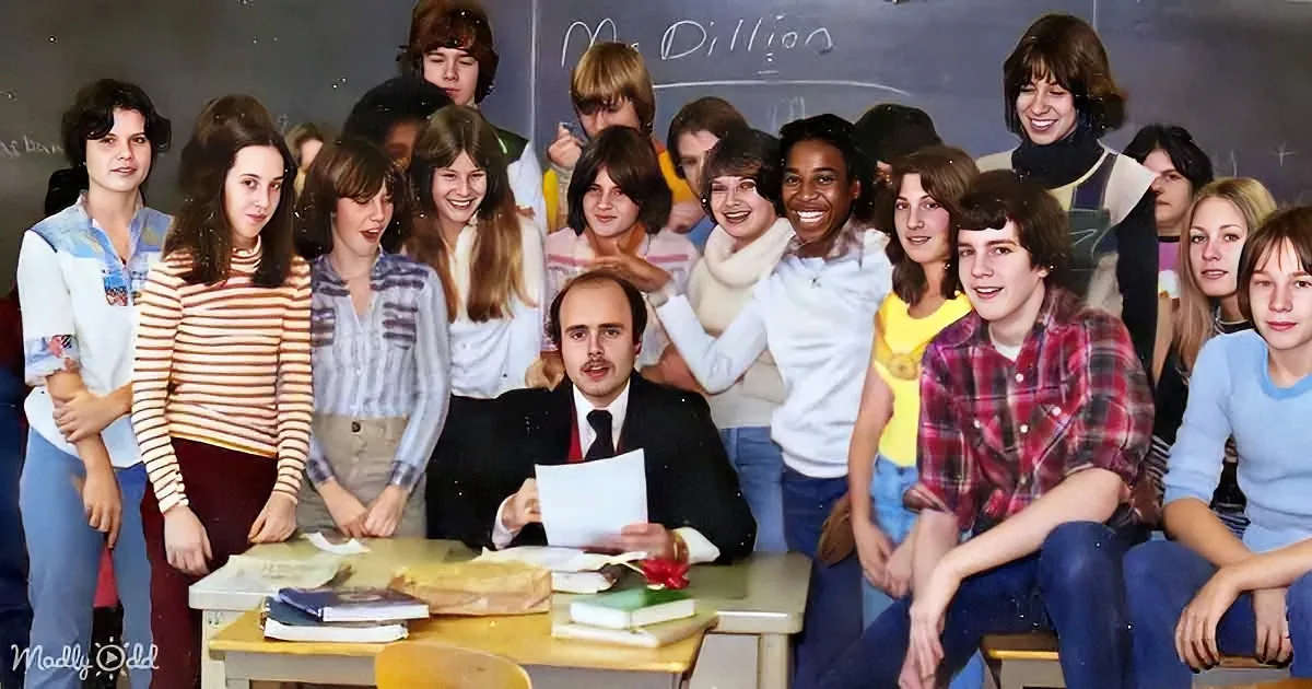 Teacher in suit sits at desk with students in colorful clothing, smiling in a classroom.