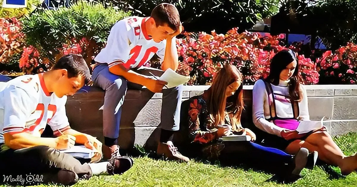 A young man in a white jersey sits on a bench, reading a book, with others studying nearby in a sunny park.
