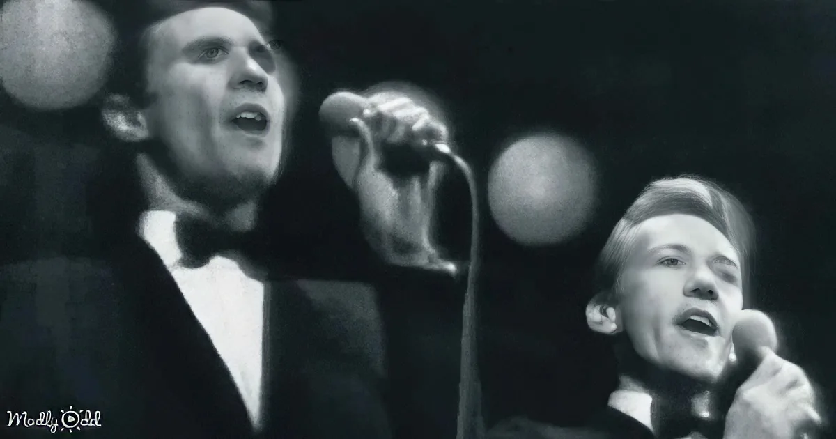 A man in a tuxedo sings into a microphone, with another person partially visible behind him.