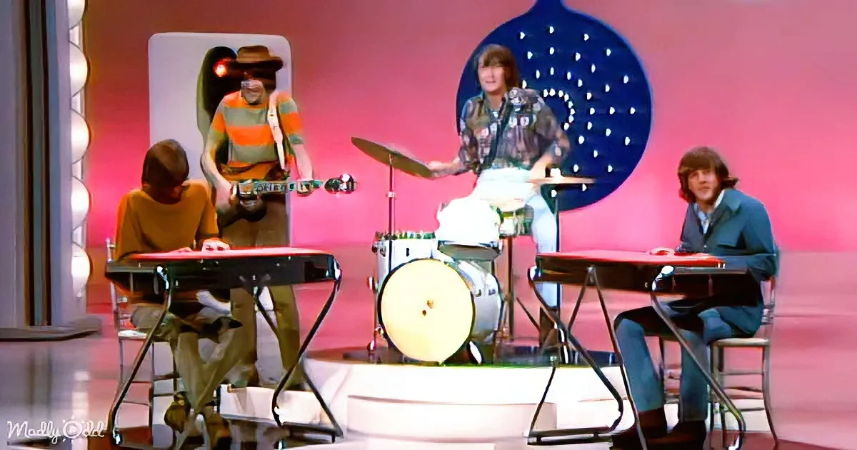 A band performs on stage with a pink backdrop, featuring a drummer, guitarist, keyboardist, and another keyboardist.