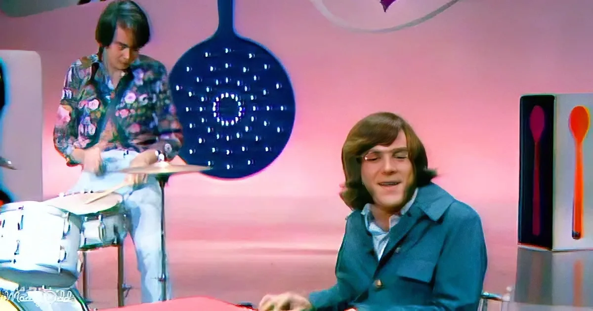 A man in a blue jacket sits smiling, while another plays drums in a colorful, retro-styled setting.