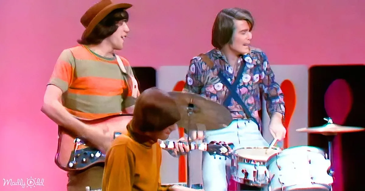 A man in a striped shirt plays guitar while another in a floral shirt plays drums, both on stage with a pink background.
