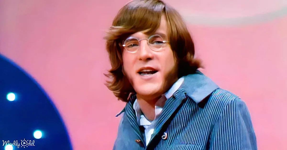 A person with shoulder-length brown hair and glasses, wearing a blue striped shirt, is speaking against a pink background.