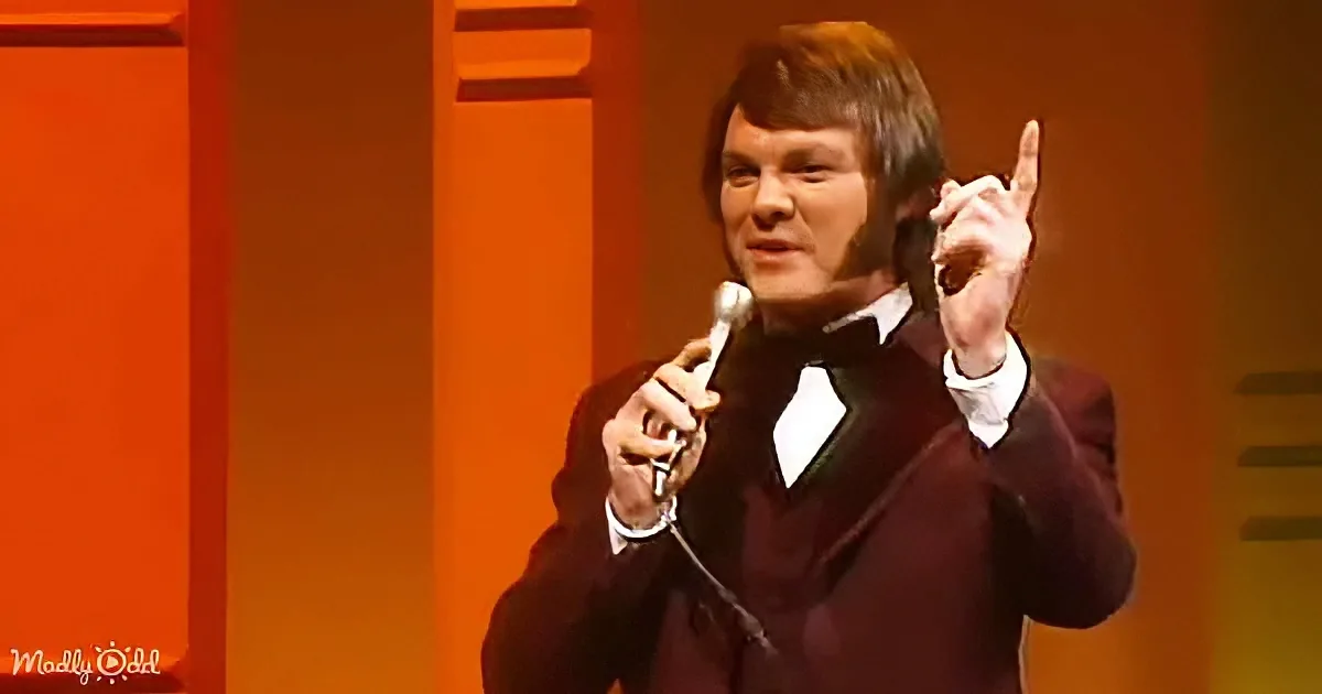 A man in a dark suit and bow tie holds a microphone, gesturing with his right hand.