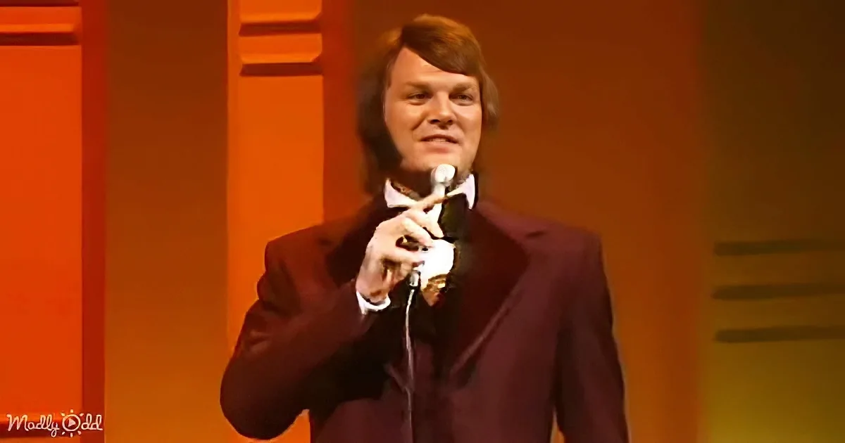 A man in a dark suit holds a microphone on stage, smiling.