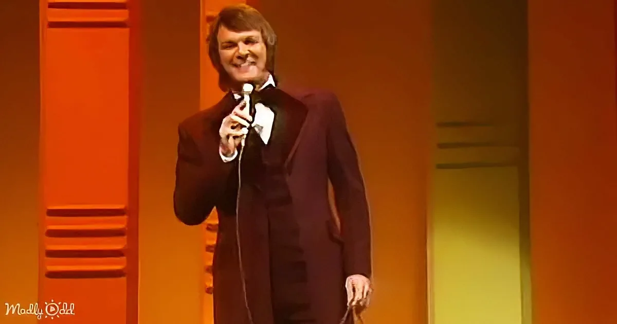 A man in a dark suit holds a microphone, smiling on stage with orange and yellow lighting.