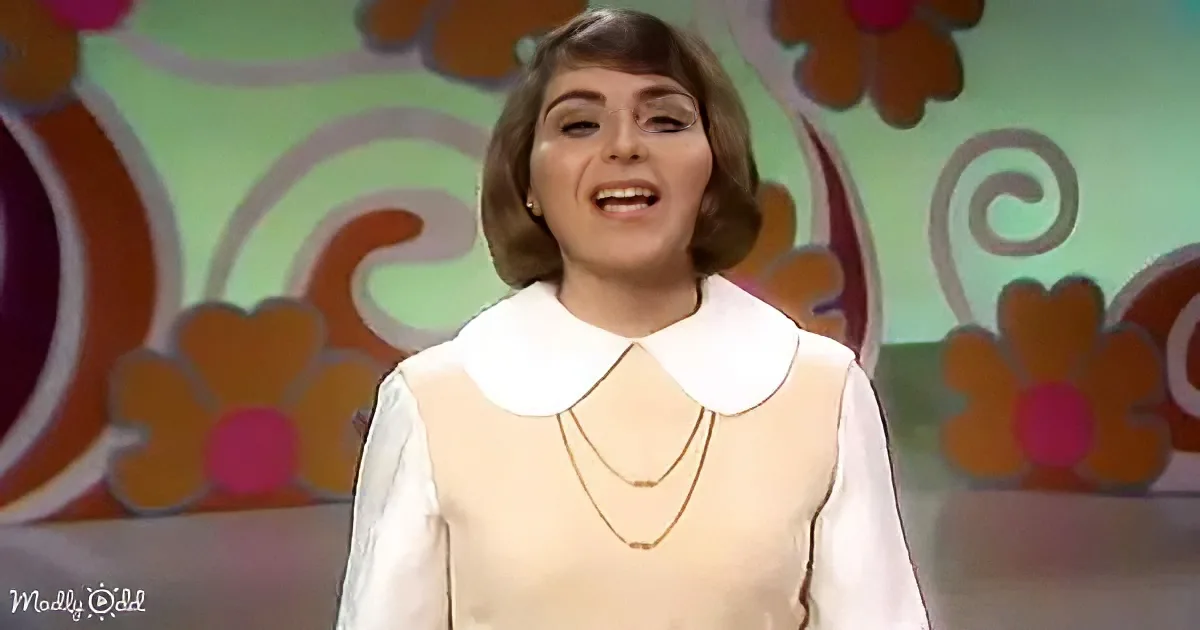 A woman with short brown hair, wearing a white blouse with a Peter Pan collar and layered necklaces, stands against a...