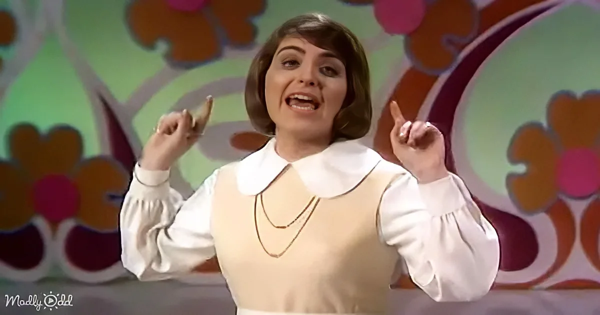 A woman in a white blouse with a Peter Pan collar and layered necklaces, pointing upwards with both hands.