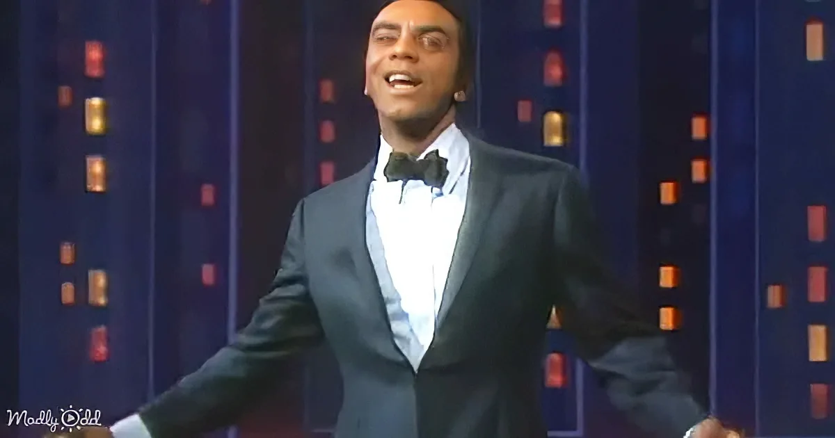 A man in a tuxedo and bow tie is smiling and gesturing on stage with a cityscape background.