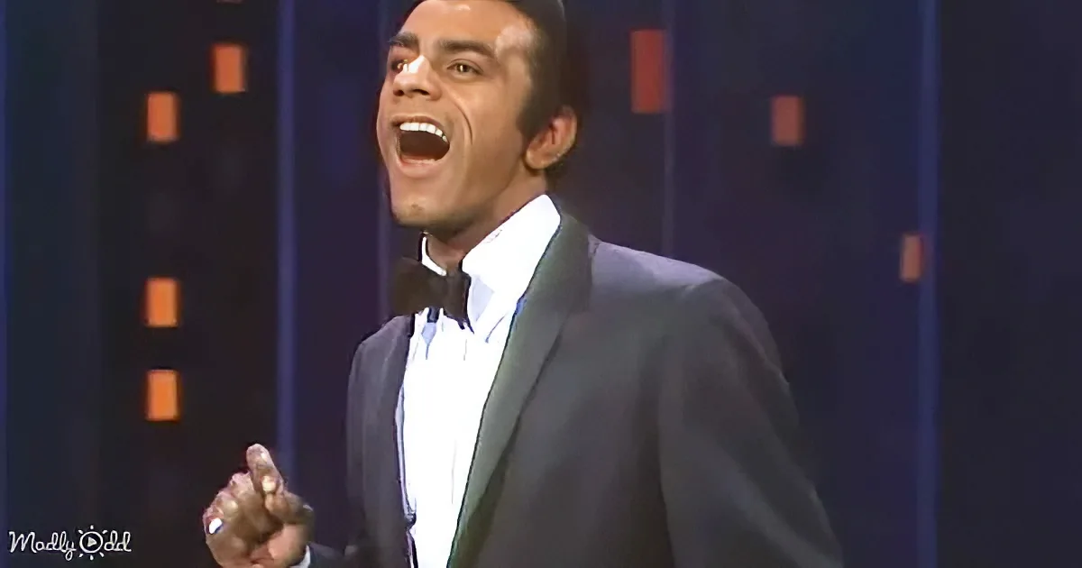 A man in a tuxedo is singing passionately with his eyes closed and hand raised.