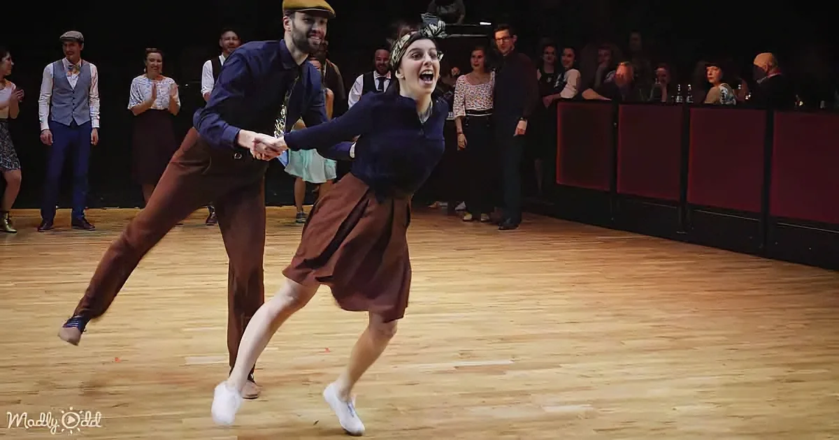 A couple dances energetically on a wooden floor, surrounded by an audience in a dimly lit venue.