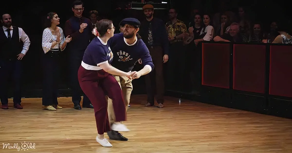 A couple in vintage attire dances on a wooden floor, surrounded by an audience watching.