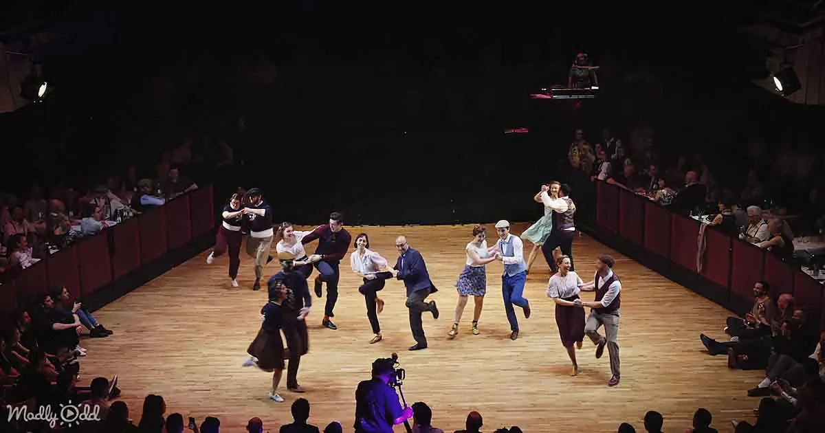 A lively dance performance with multiple couples on a wooden stage, surrounded by an audience.