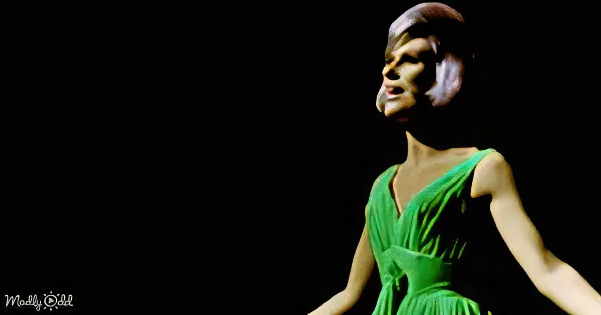 A person in a green dress with a dark background, arms outstretched.