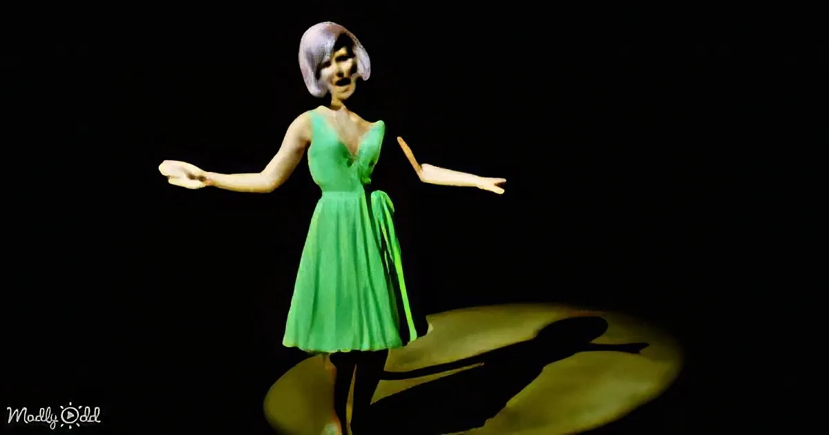 A woman in a green dress stands with arms outstretched, illuminated against a dark background.