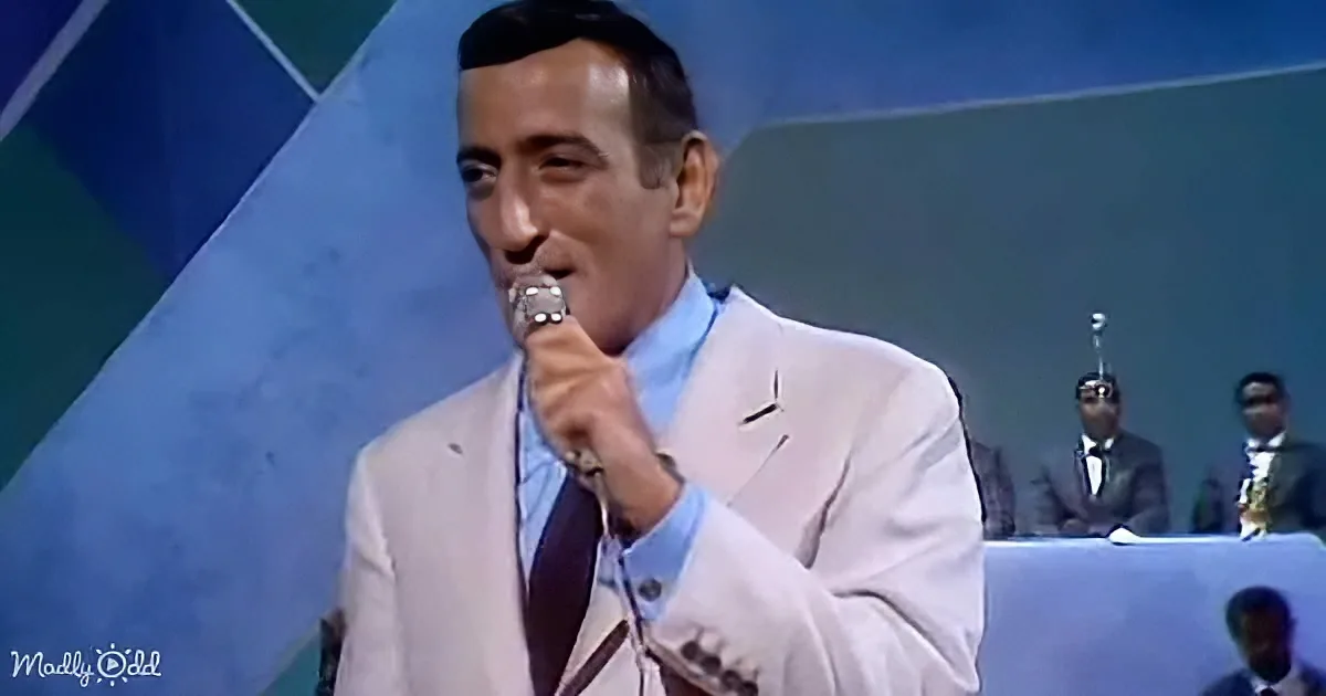 A man in a light suit holds a microphone, singing on stage with a band in the background.