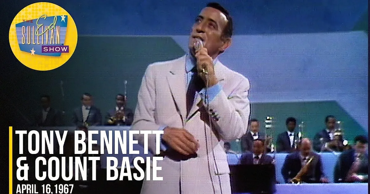A man in a striped suit holds a microphone, singing with a smile, against a geometric background.