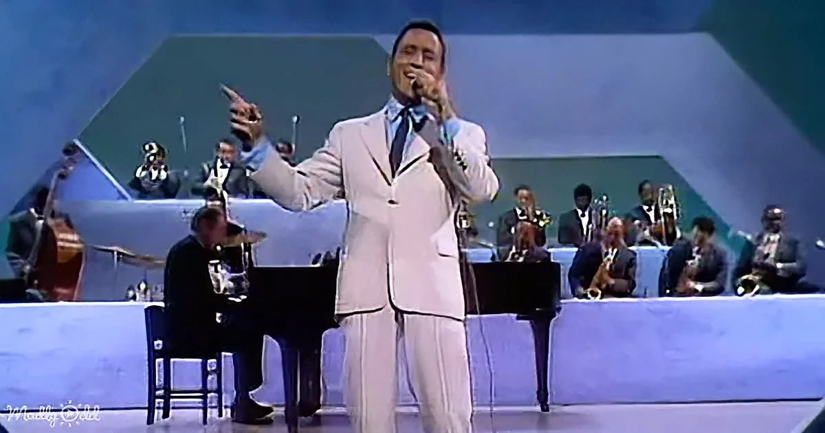 A man in a white suit sings on stage with a band in the background.