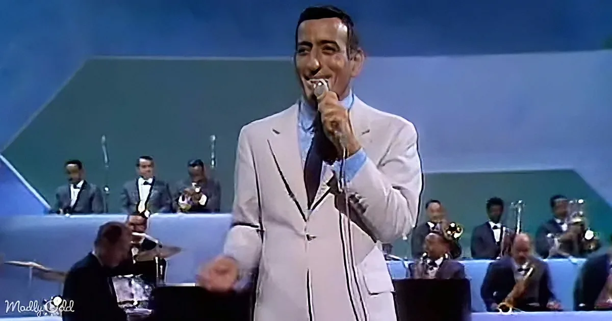 A man in a white suit sings into a microphone, smiling, with a band in the background.