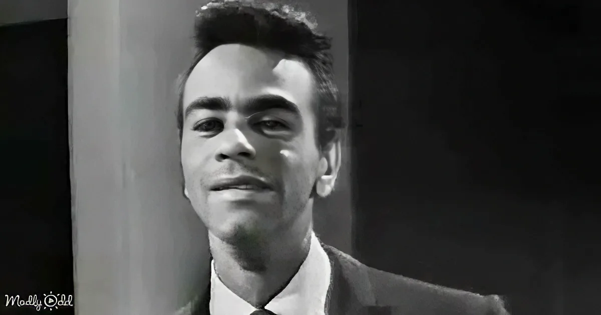 A man in a suit and tie smiles slightly, looking to the side, with a dark background.