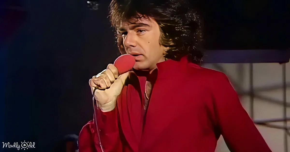 A man in a red outfit sings into a red microphone, with a dark background and a faint watermark.