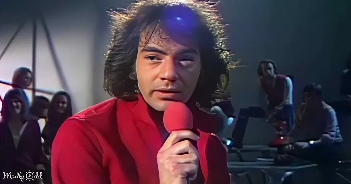 A person in a red outfit holds a microphone, surrounded by people in a dimly lit setting.