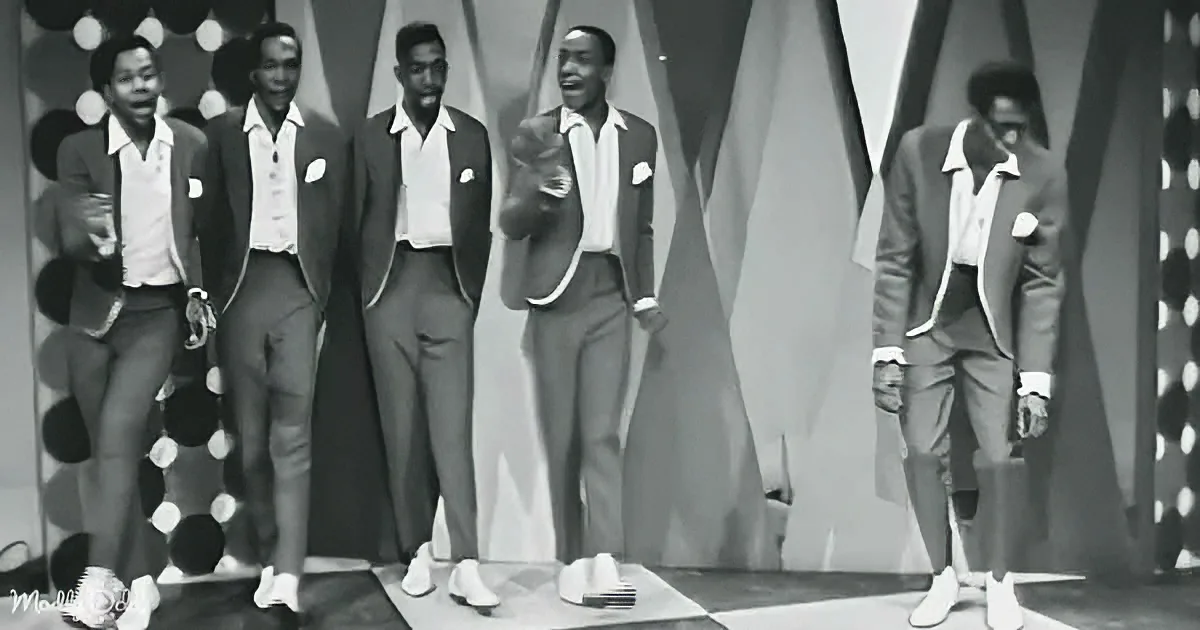 A group of five men in suits and ties perform on a stage with a patterned backdrop.