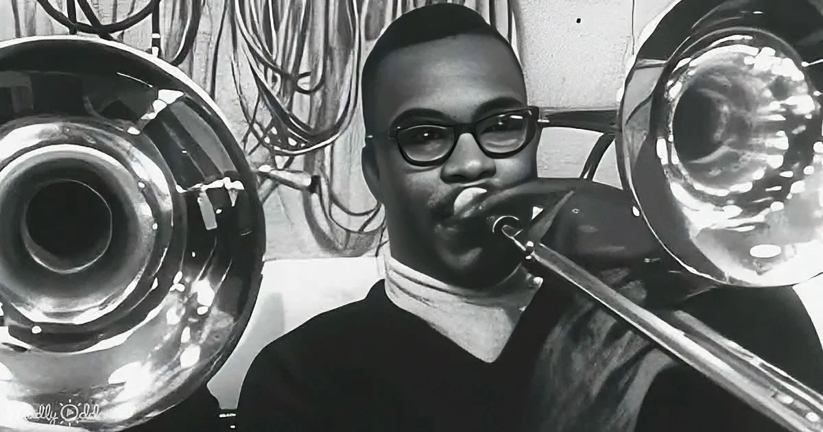 A person wearing glasses plays a trombone, surrounded by other brass instruments.