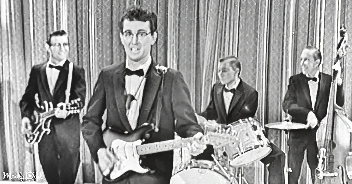 A man in a suit plays a guitar, surrounded by band members in formal attire, on a stage with a curtain backdrop.