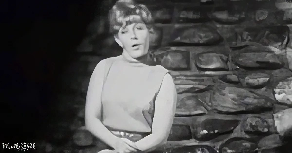 A woman in a turtleneck sits in front of a stone wall, hands clasped, in a black-and-white image.