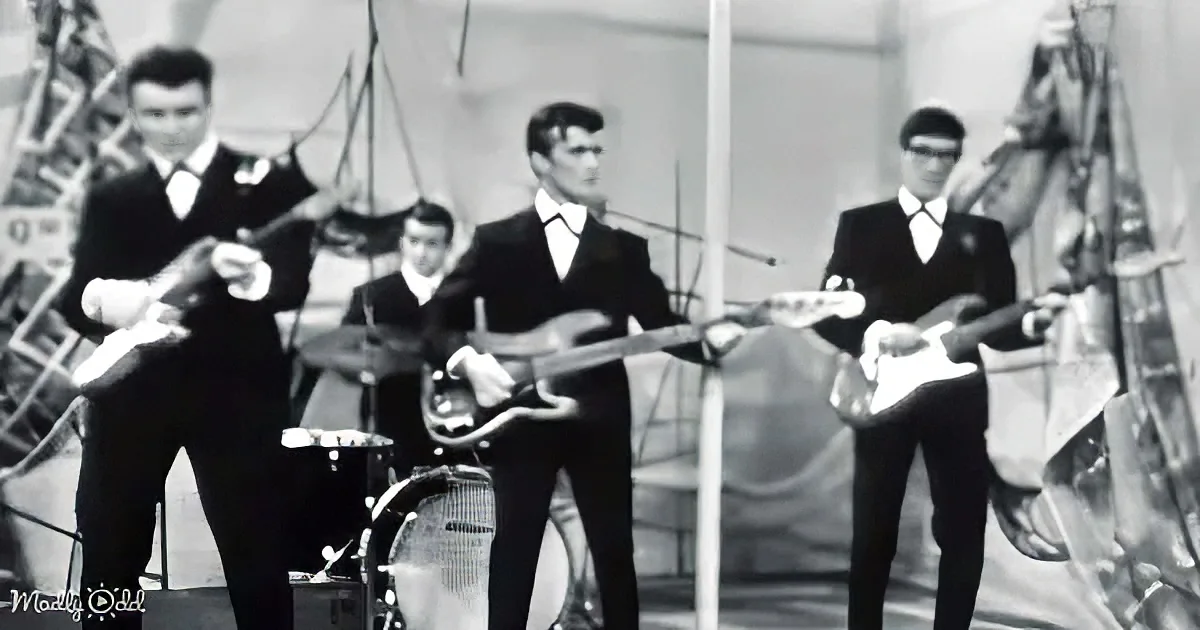 A group of musicians in suits play guitars and drums on stage, with a backdrop of ropes and flags.