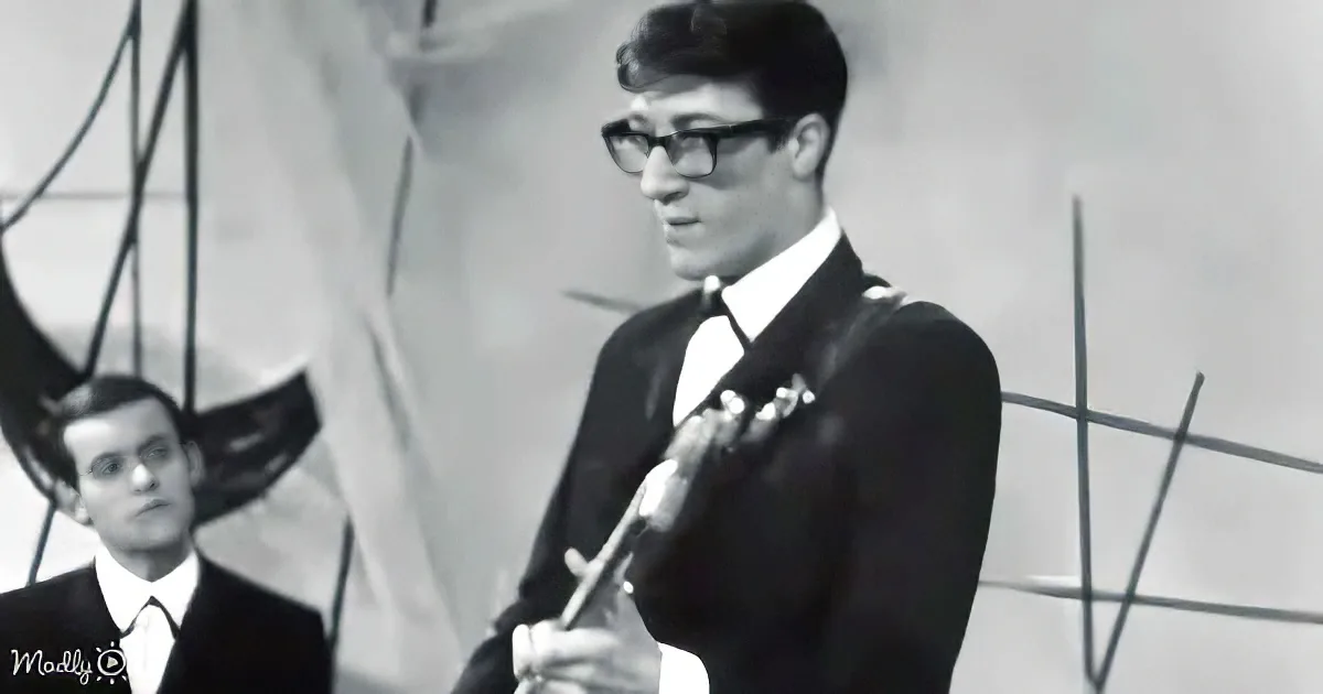 A man in a suit and glasses plays a guitar, with another man in the background.