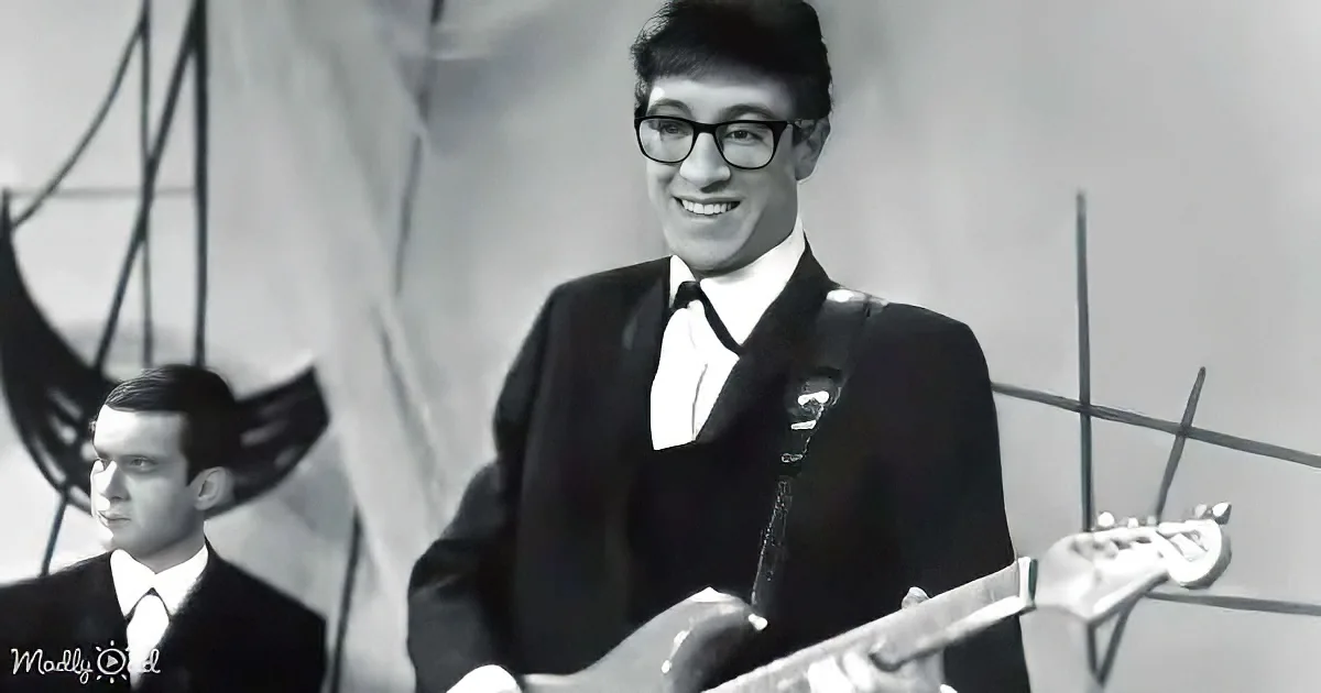 A man in a suit and tie smiles while holding a guitar, with another person in the background.