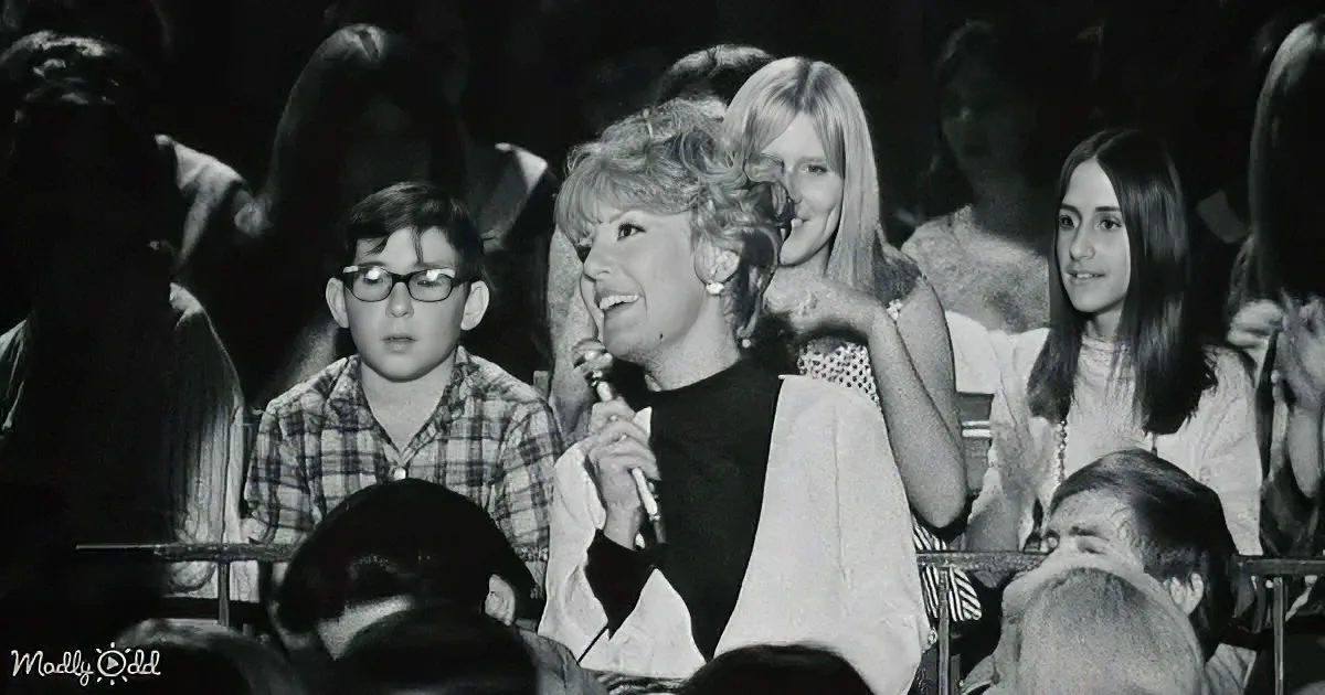 A woman with a microphone smiles amidst a crowd of people in a dimly lit setting.