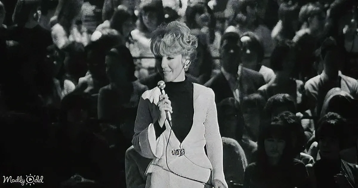 A woman in a white suit holds a microphone, standing in front of a crowd, smiling.