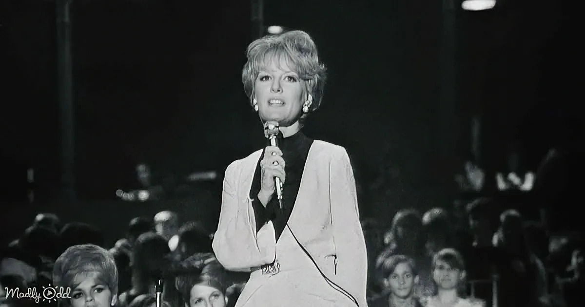 A woman in a white suit holds a microphone on stage, with a crowd in the background.