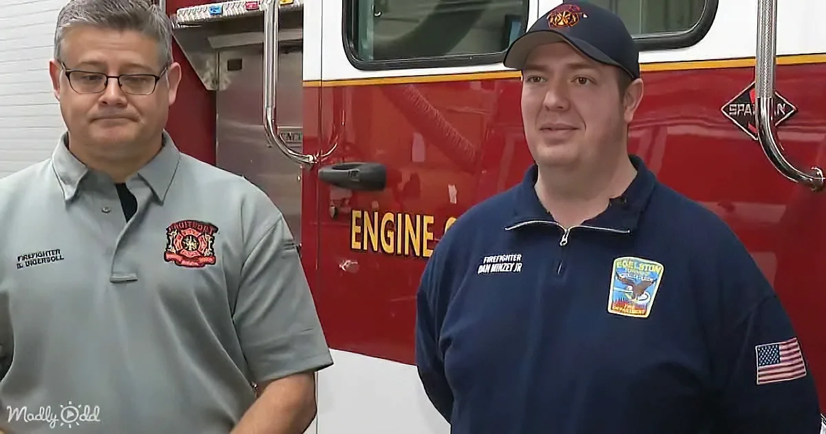 Two firefighters in uniform stand in front of a red fire truck, smiling.