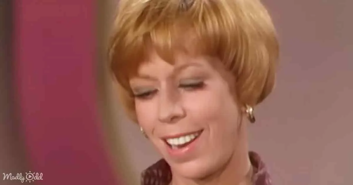 A smiling woman with short blonde hair, wearing earrings, and a maroon top, set against a pink and beige background.