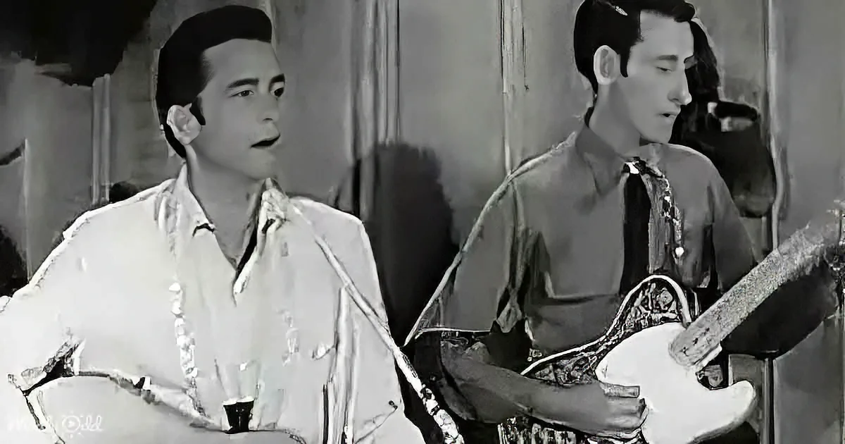 A man in a white shirt sits, while another plays a guitar, both in a dimly lit setting.